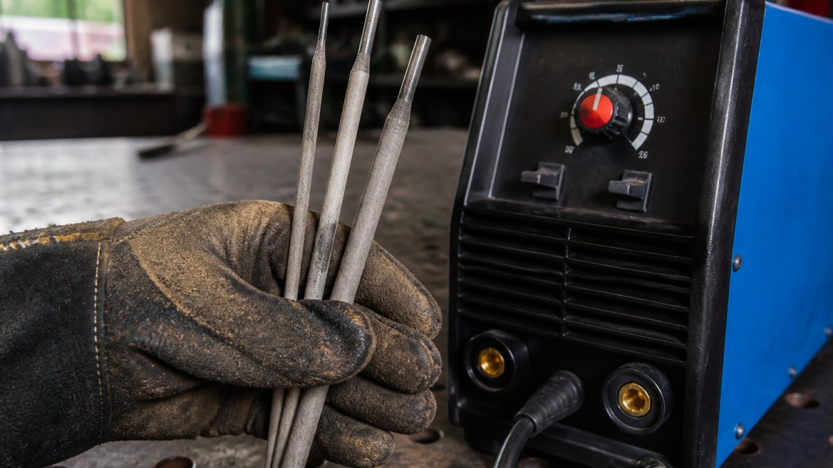 Professional welder's gloved hand holding different electrode rods (1.6mm, 2.5mm, 3.2mm sizes) positioned next to a welding machine control panel showing amperage dial and settings in workshop lighting